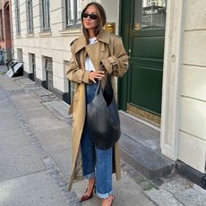 fashion influencer Felicia Akerstrom poses on a sidewalk in Copenhagen wearing black oval sunglasses, a trench coat, white t-shirt, cuffed jeans, black leather shoulder bag, and burgundy slingback heels.