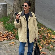 French Fashion Trends 2025: @salome.mory wears a barn jacket with blue jeans and brown boots whilst standing on a leafy street