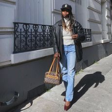 Fashion influencer @aude_julie on the street wearing a vintage-look, collared, black leather jacket with a turtleneck, blue faded jeans, burgundy heels, a Louis Vuitton handbag, and cap.