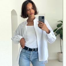 influencer Tylynn Nguyen posing for a mirror selfie wearing an open white button-down shirt, white T-shirt, black belt, and classic jeans