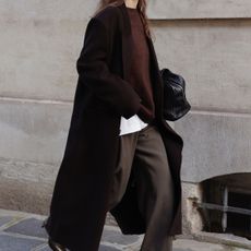 @modedamour wearing a dark, long coat, a burgundy sweater, brown pants, and a black bowler bag.