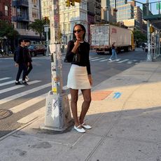 Woman poses on an NYC street wearing a white mini skirt, white The Row Mary Janes and a black fitted sweater with a crewneck t-shirt peeking out of the top.